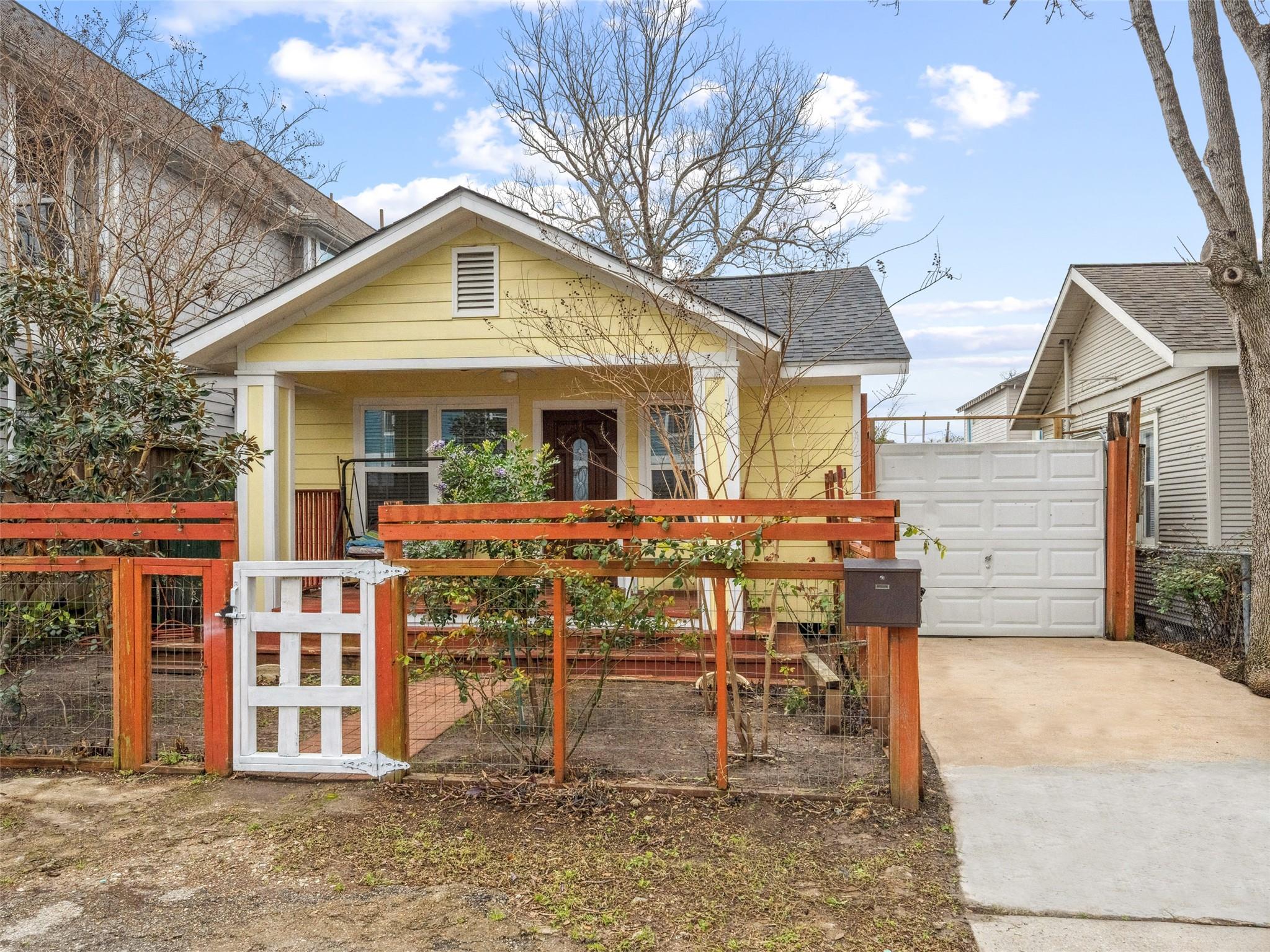 Yellow single-story house with a covered porch, reddish wooden fence, white gate, and driveway to a detached garage.