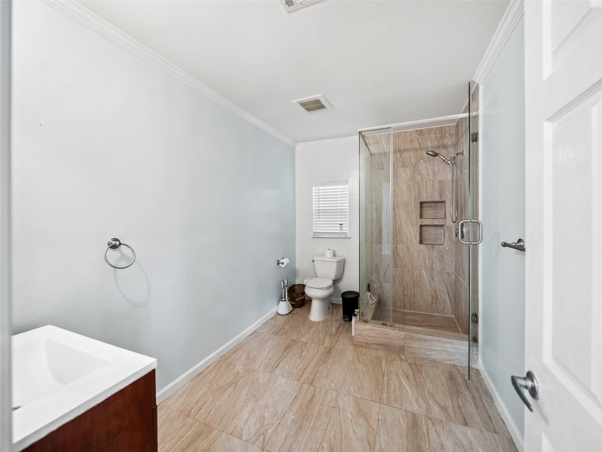 Bathroom with a glass-enclosed tiled shower, beige tile floors, and a window with white blinds.