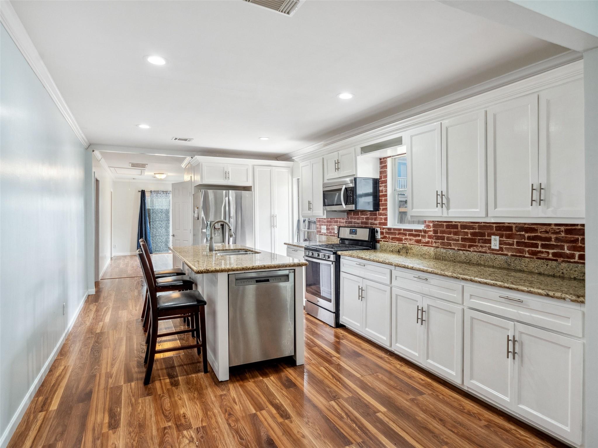 Kitchen with white cabinets, red brick backsplash, granite island, stainless steel appliances, and wood floors.