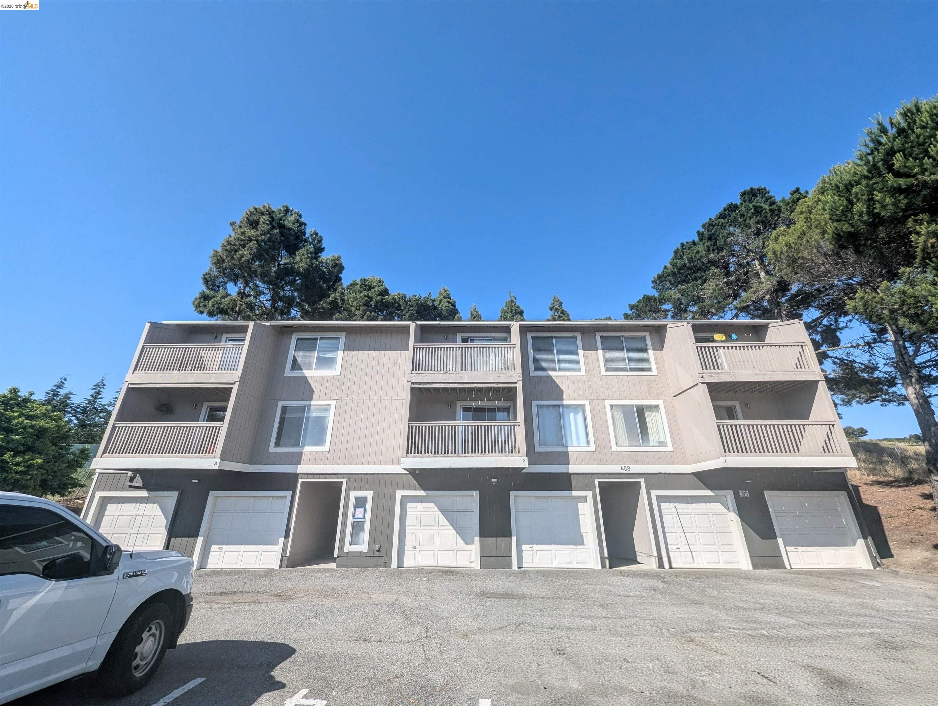 Three-story gray residential building with balconies and ground-level garage doors beneath tall trees.