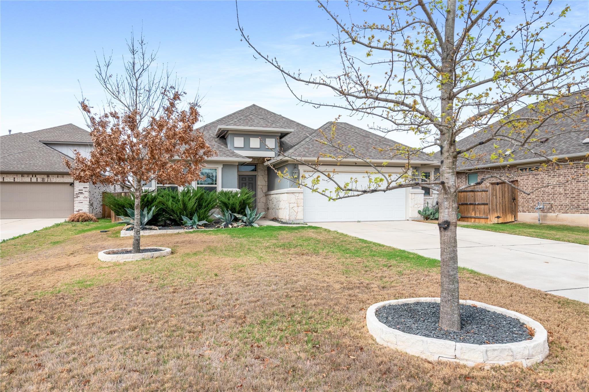 Gray single-story home with a stone-accented entry, white garage door, and two bare front-yard trees.