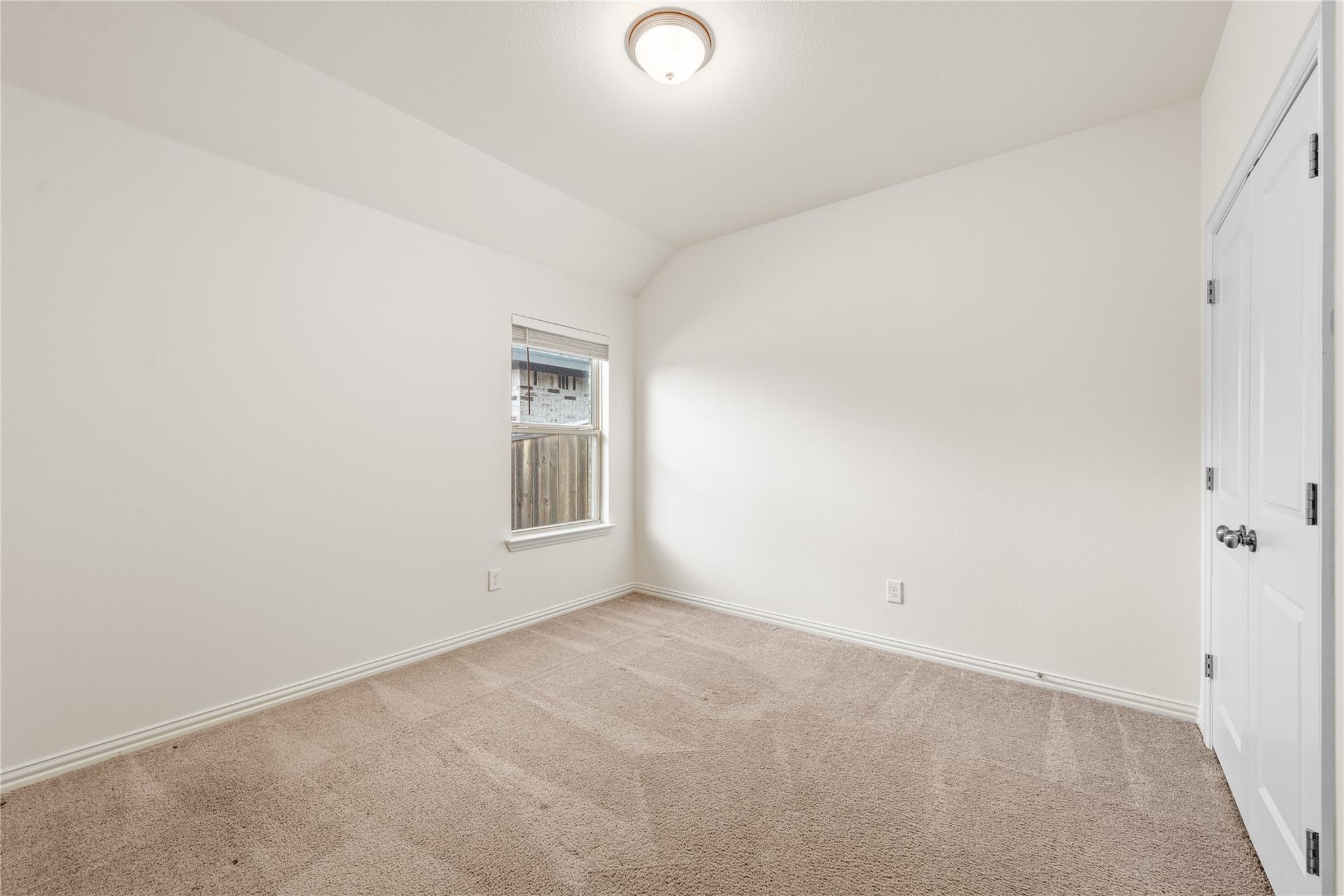 Empty bedroom with beige carpet, a window with blinds, and white closet doors.