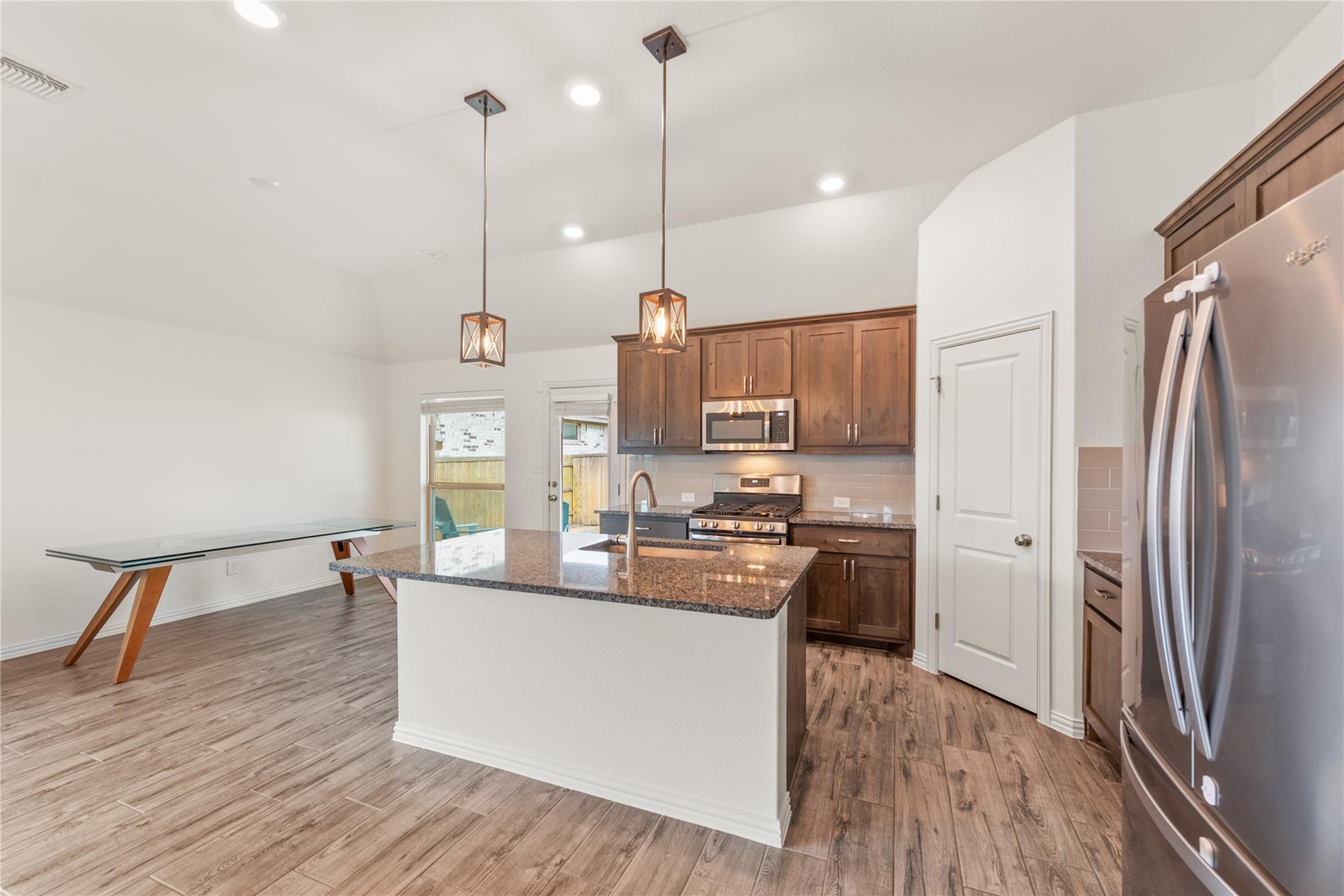 Open kitchen with dark wood cabinets, stainless steel appliances, and a granite island beside a glass dining table.