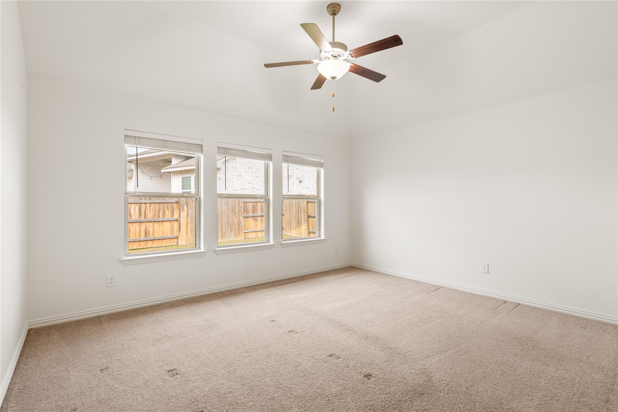 Empty bedroom with beige carpet, three windows, and a ceiling fan with light.