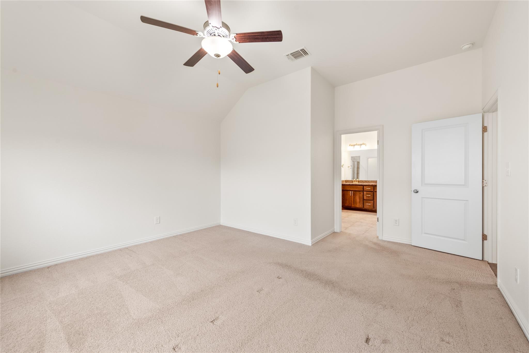 Empty carpeted bedroom with a ceiling fan and an open doorway to a bathroom vanity.