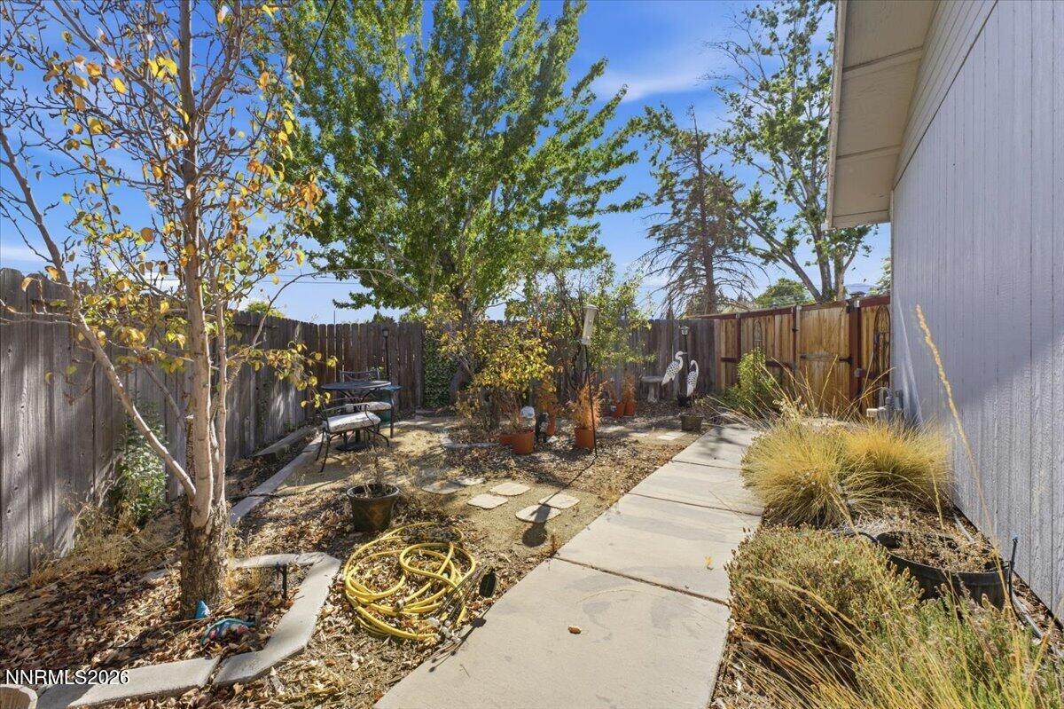 Fenced backyard with a concrete walkway, mature trees, and patio seating.