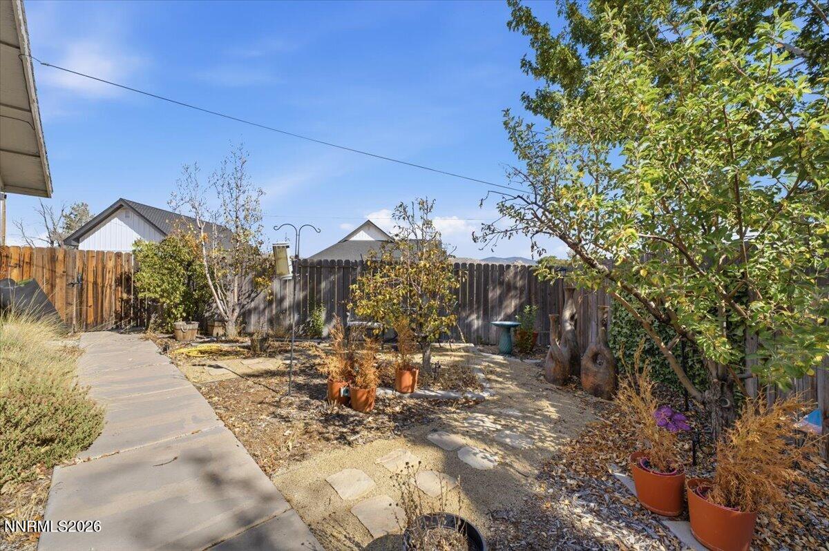 Fenced backyard with a paved walkway, garden beds, potted plants, and distant mountain ridges.