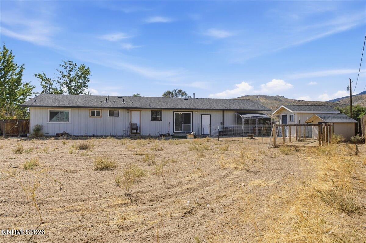 Rear of a single-story home with a dry fenced yard, outbuildings, and distant hills.