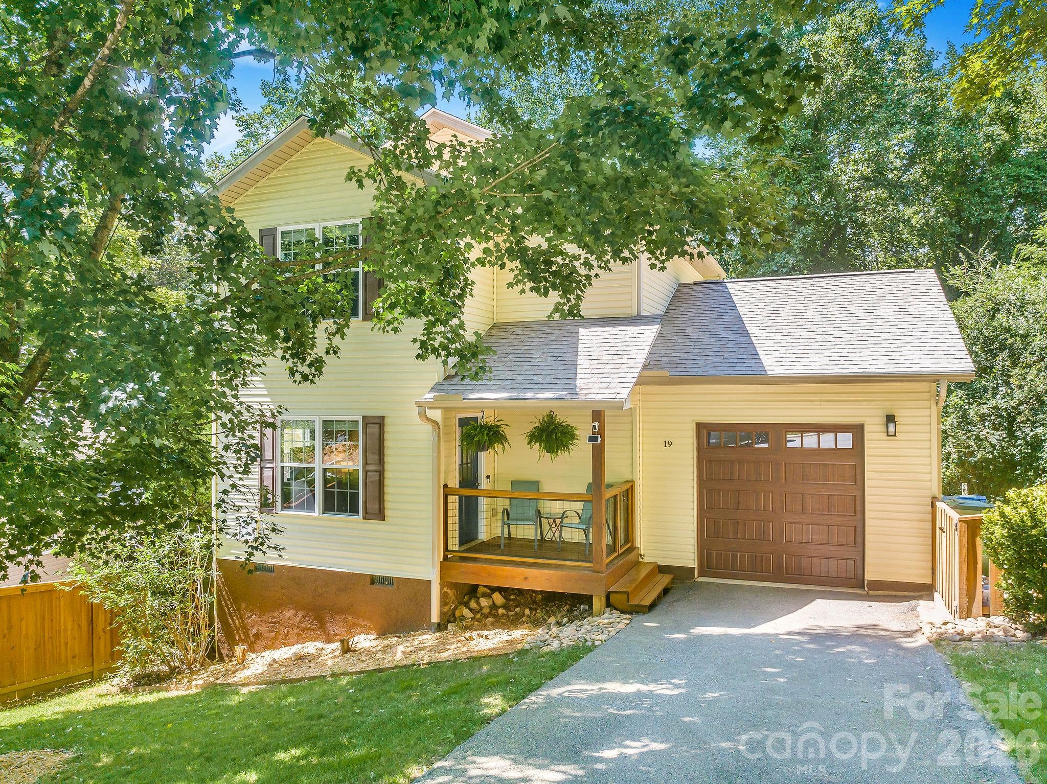 Pale yellow two-story house with a covered porch, attached garage, and leafy trees.