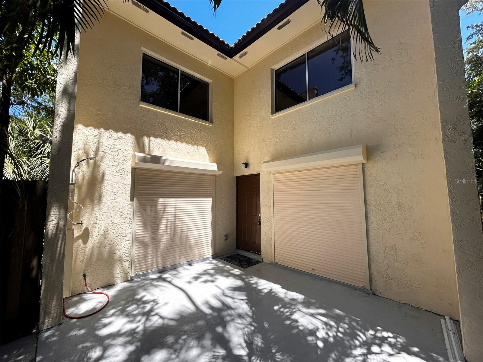 Paved courtyard beside a beige stucco home, with roll-down shutters and palm-tree shadows.