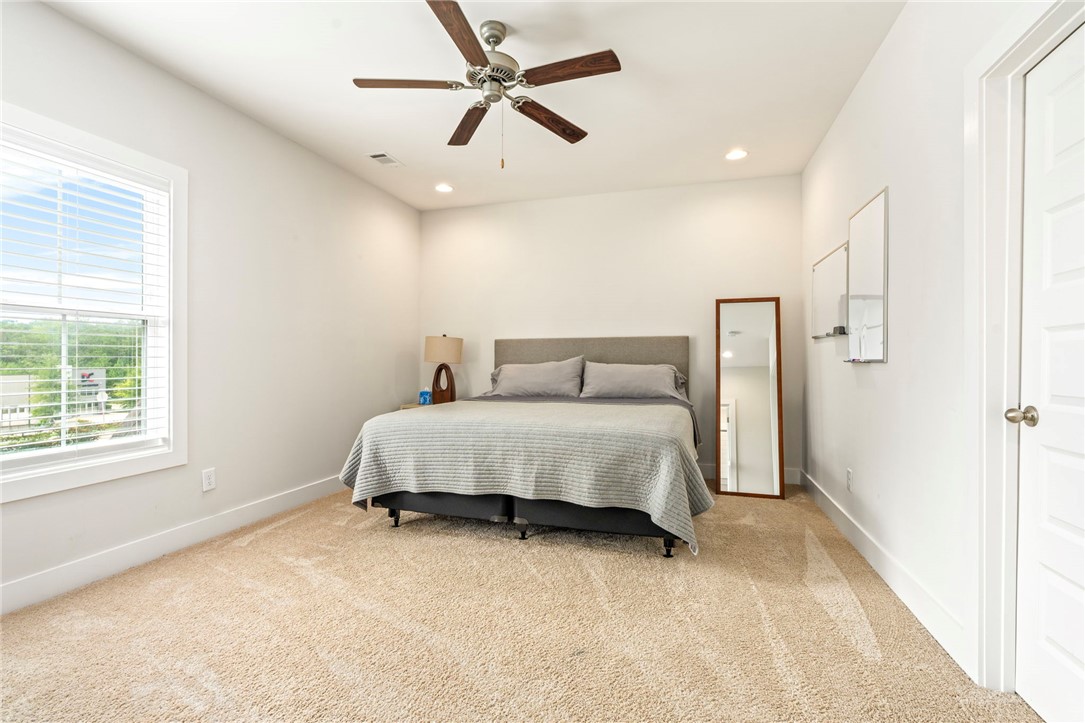 Furnished bedroom with a king bed, beige carpet, large window, and ceiling fan.