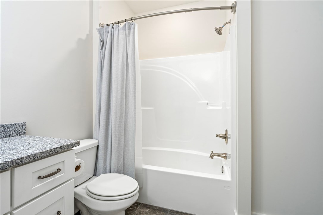 Bathroom with a white tub and shower, gray curtain, toilet, and granite-topped vanity.