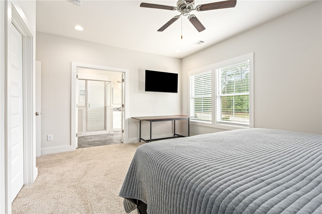 Bedroom with a gray quilted bed, bright windows, ceiling fan, and open doorway to an attached bathroom.