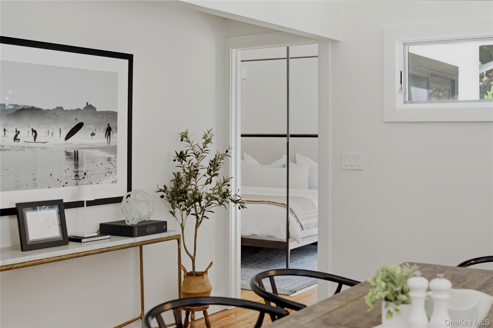 Staged dining area with black chairs and a wooden table, looking through to a white bedroom.