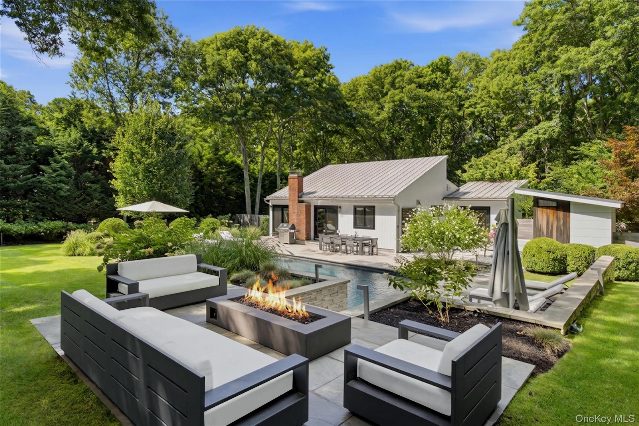 Modern home with a pool, patio lounge seating, and fire pit in a wooded backyard.