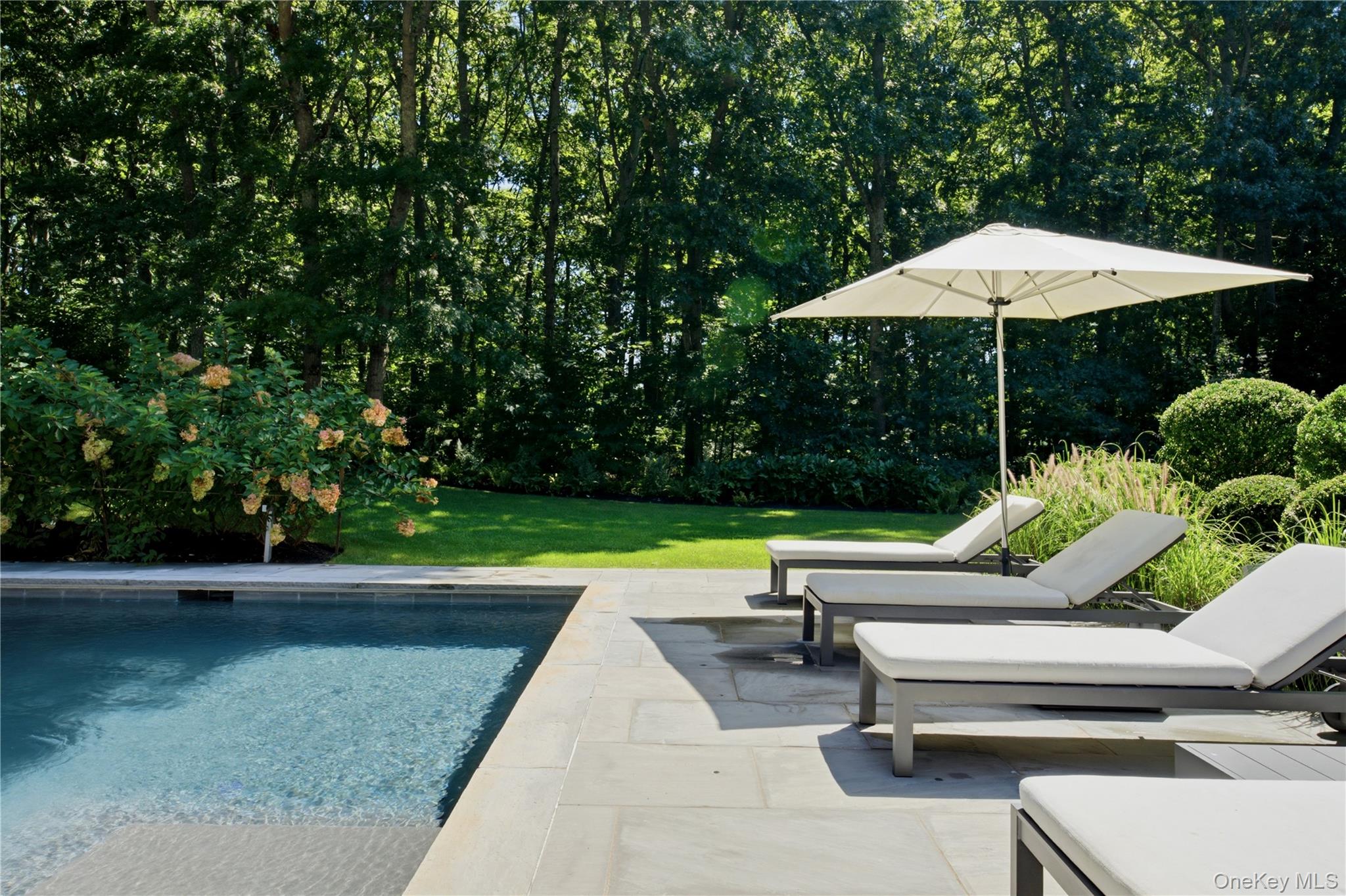 Swimming pool beside a stone patio with white loungers and a large umbrella.