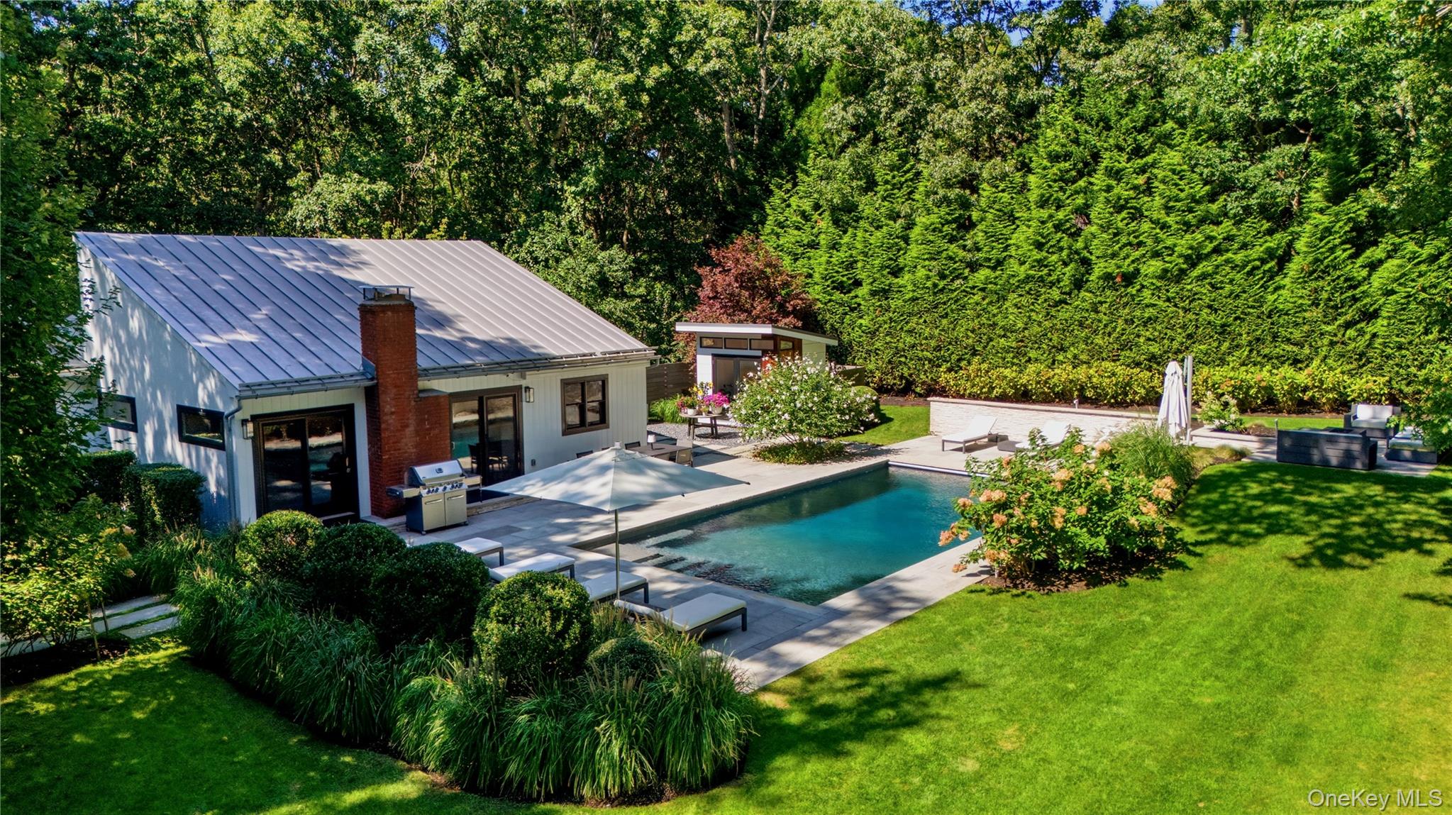 Modern home and landscaped backyard with a rectangular pool, stone patio, and tall privacy hedges.