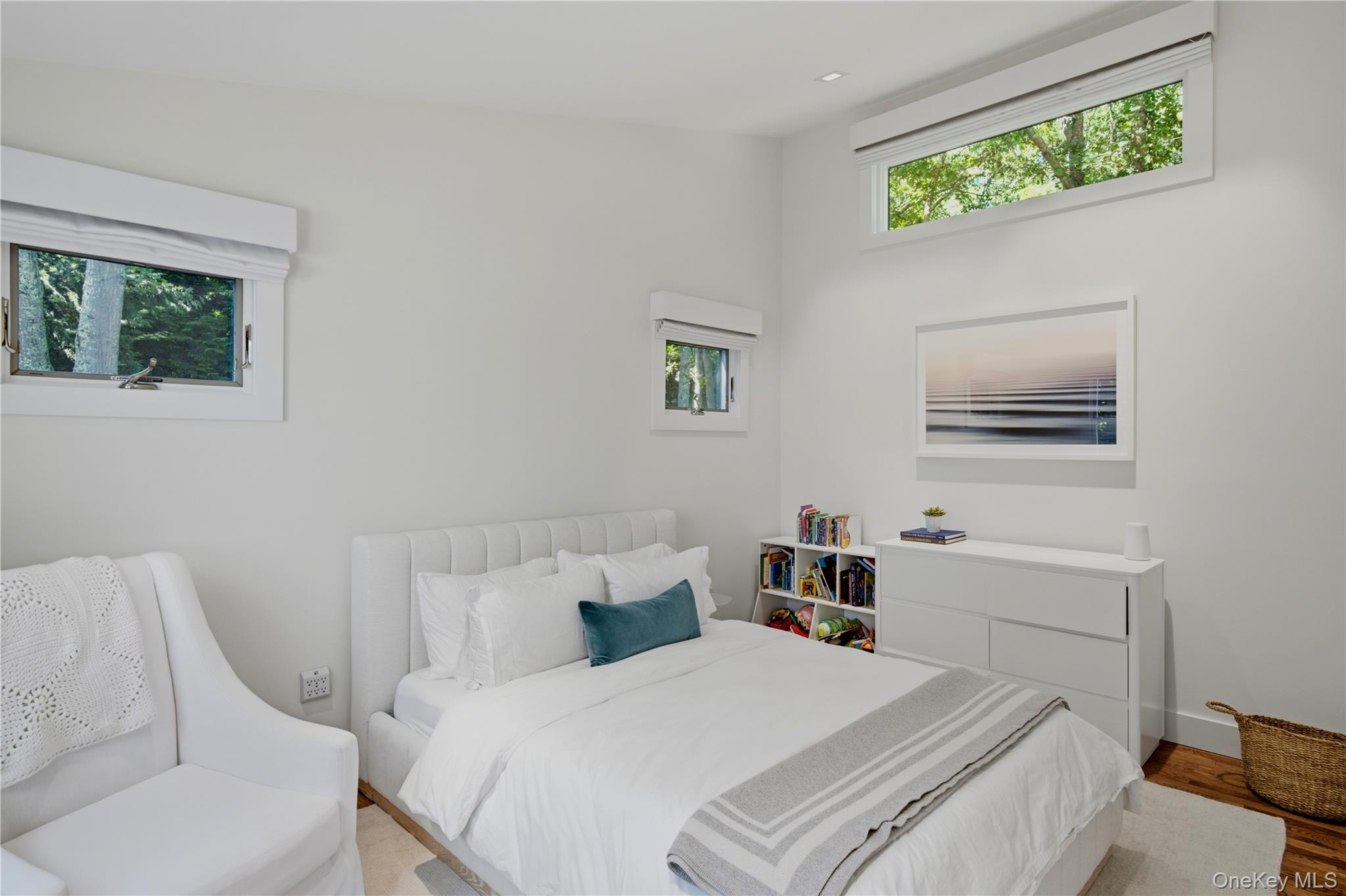 Bright white bedroom with a bed, upholstered chair, and three high windows overlooking trees.
