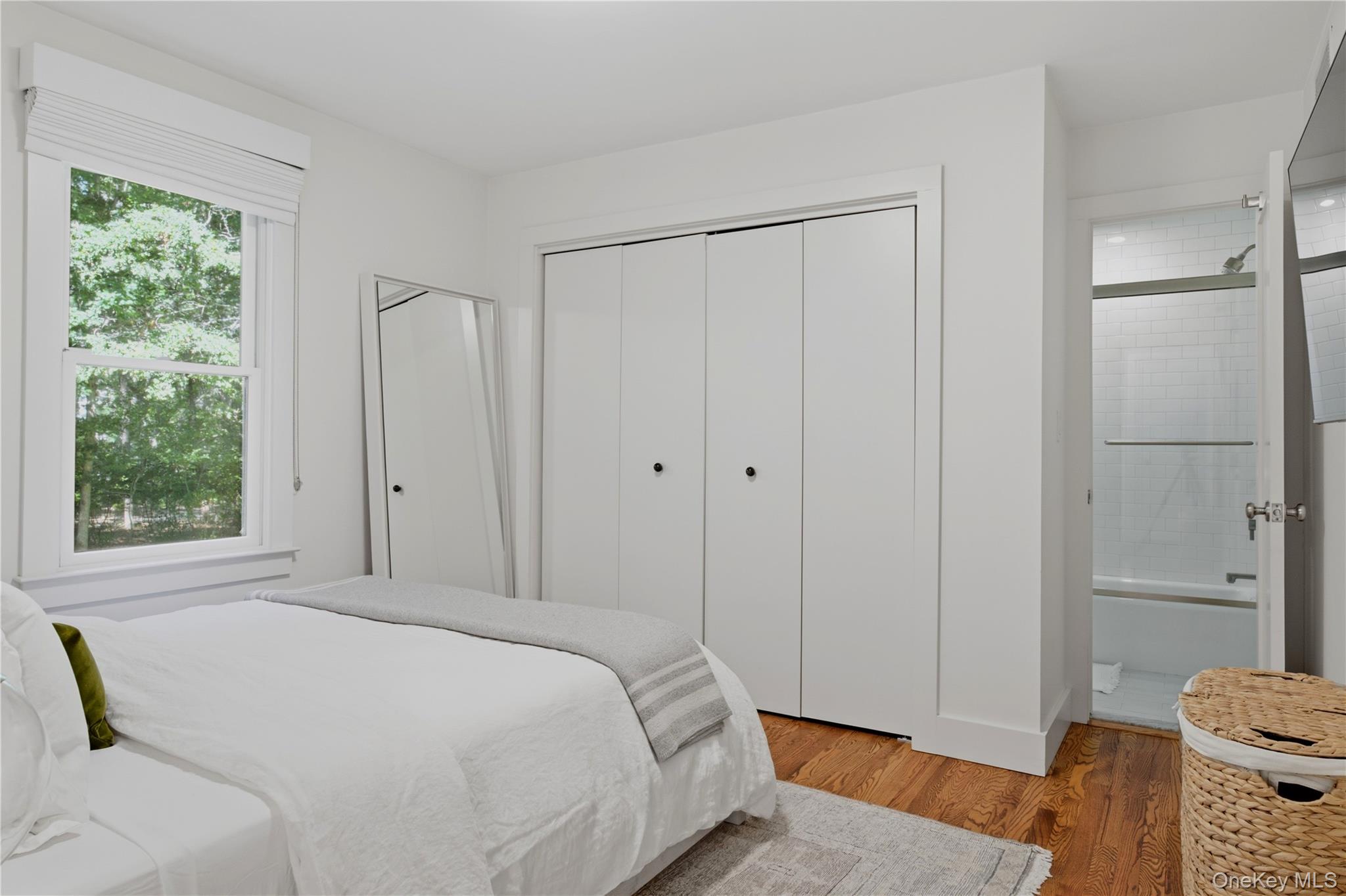 Bright bedroom with white bedding, wood floors, and an open doorway to a tiled bathroom.