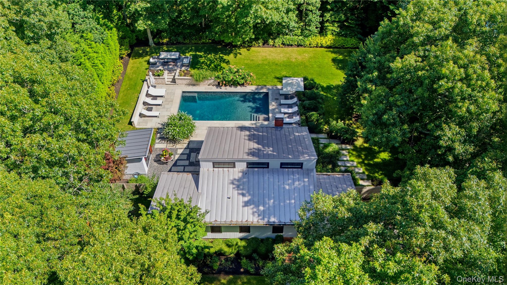 Aerial view of a modern home with a rectangular pool, stone patio, and tree-lined lawn.