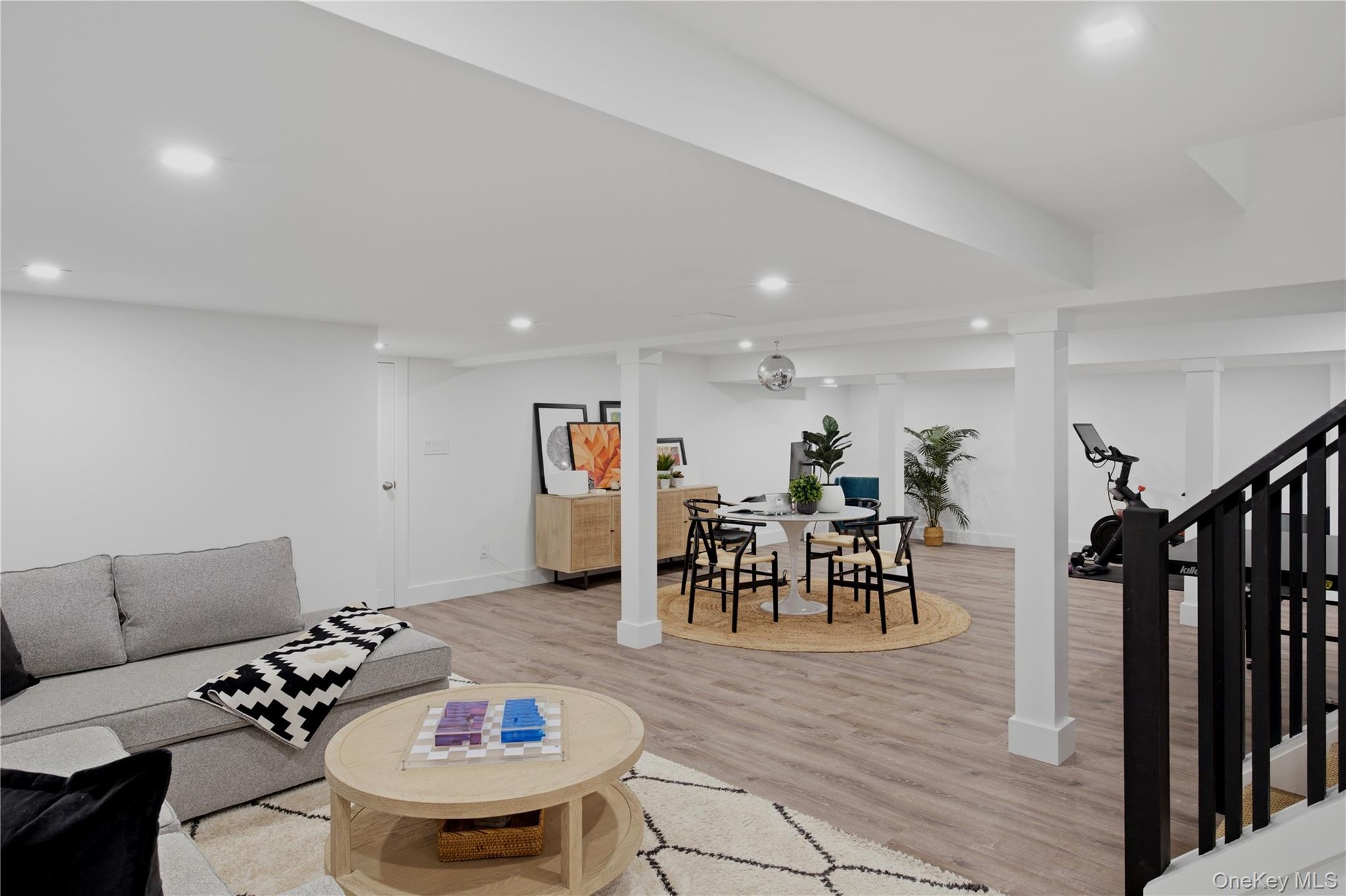 Furnished lower-level living area with a gray sectional, dining table, exercise bike, and black stair railing.