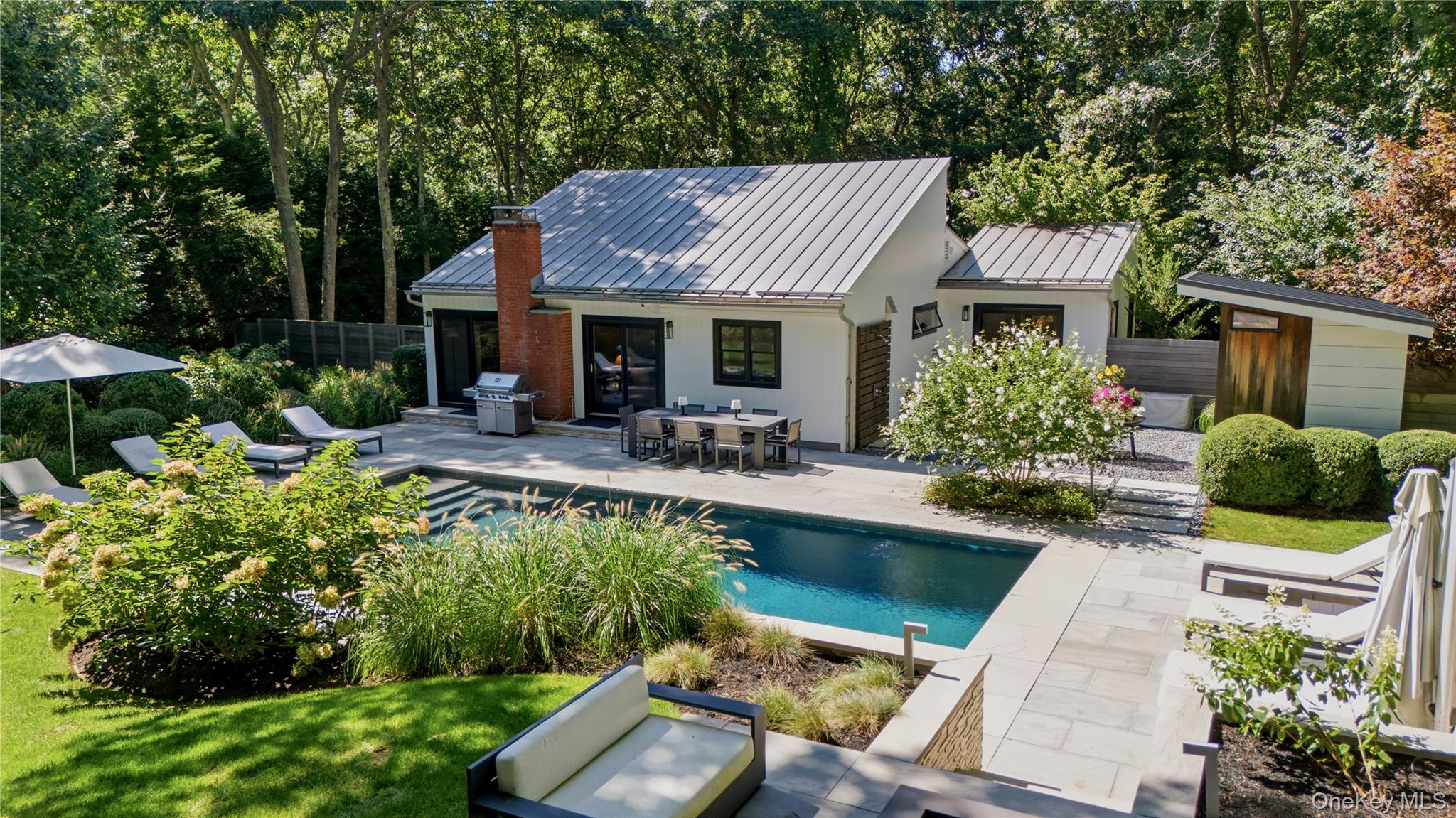 Modern white home with a metal roof, swimming pool, and furnished stone patio amid mature trees.