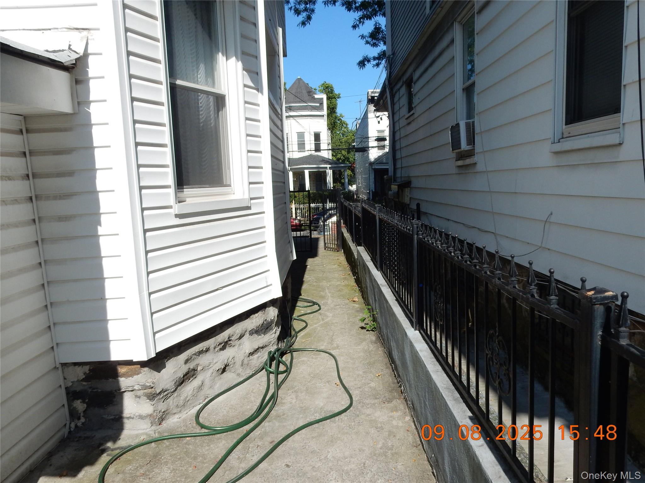 Narrow concrete side passage between houses, with a black metal fence and green garden hose.
