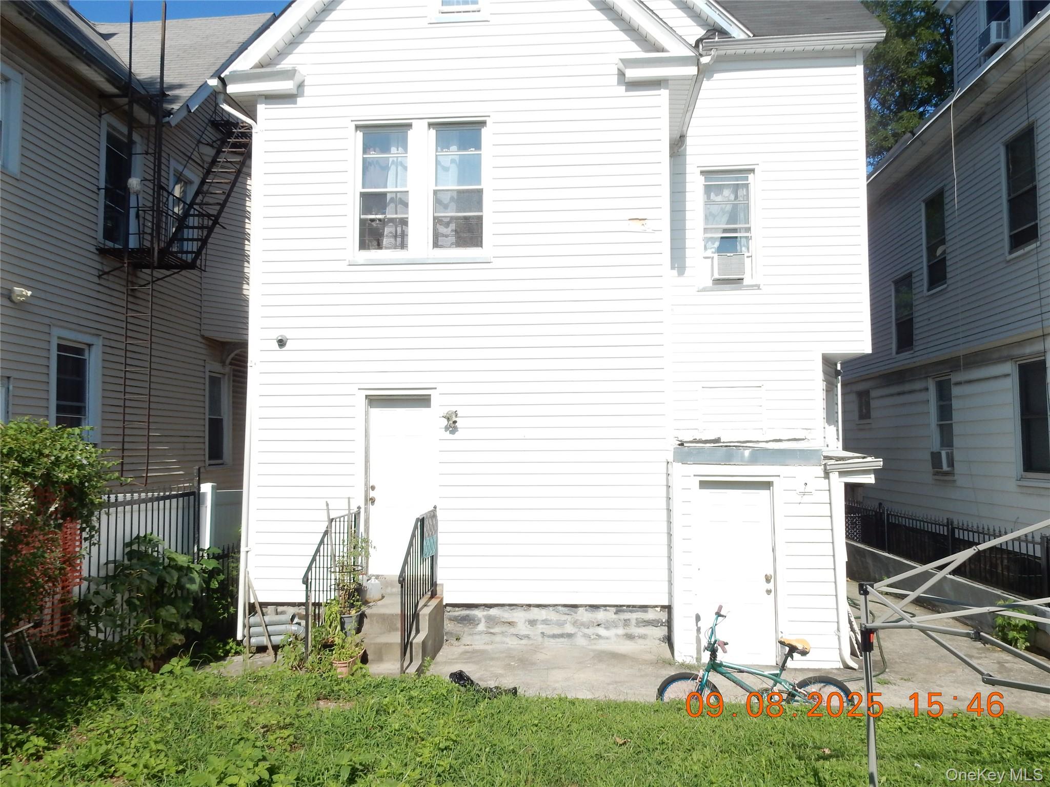 Rear of a white-sided house with a small patio, lawn, bicycle, and neighboring homes nearby.