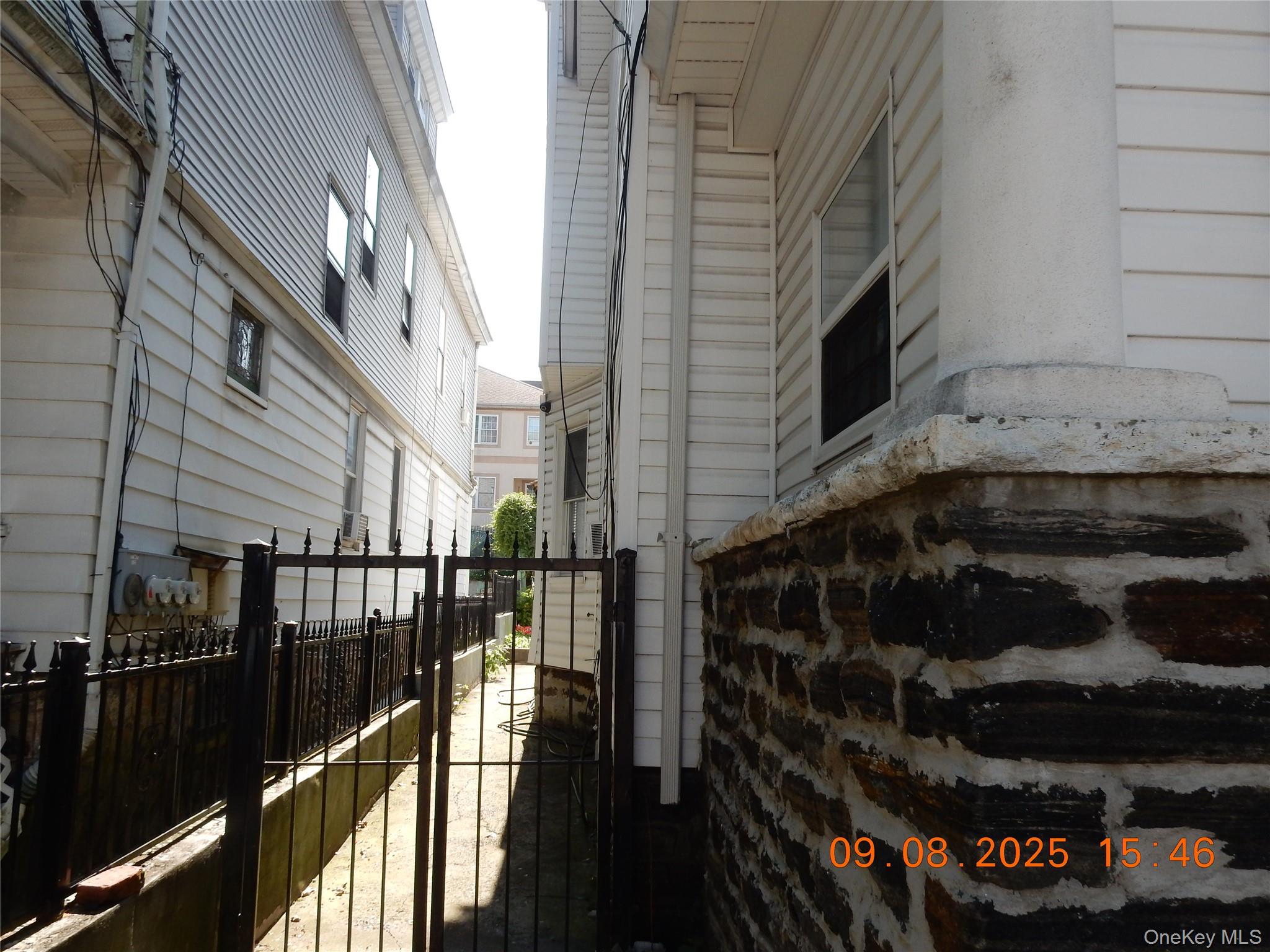 Narrow side passage between white-sided homes, with a black metal gate and stone foundation wall.