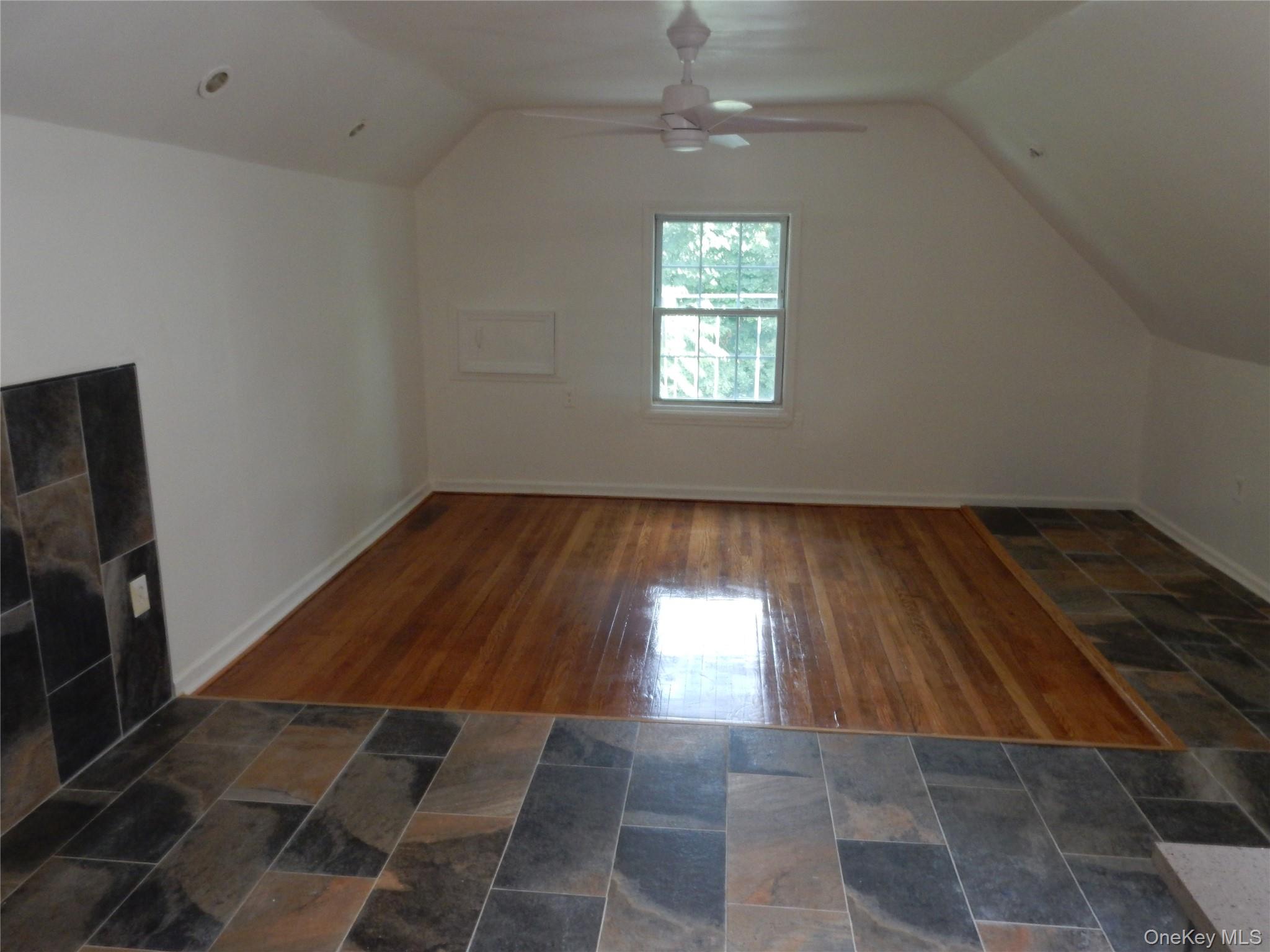Empty room with a sloped ceiling, ceiling fan, window, hardwood inset, and tile flooring.