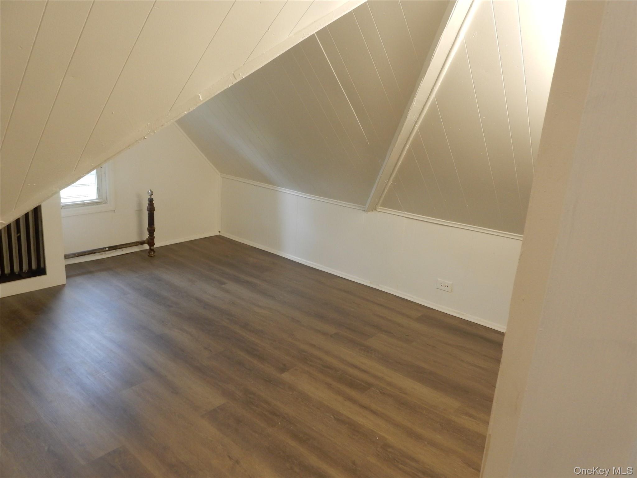 Empty attic room with sloped paneled ceilings, dark flooring, and a small window.