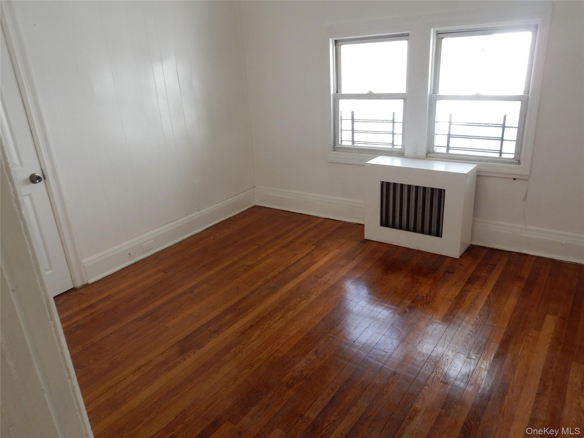 Empty room with polished hardwood floors, white paneled walls, and two windows above a radiator cover.