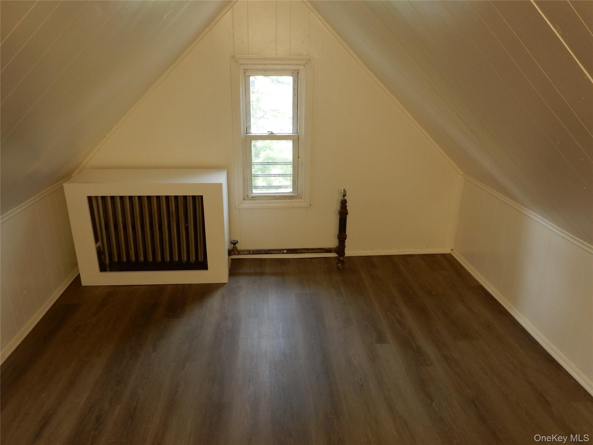 Empty attic room with sloped paneled ceiling, narrow window, and exposed radiator pipes.