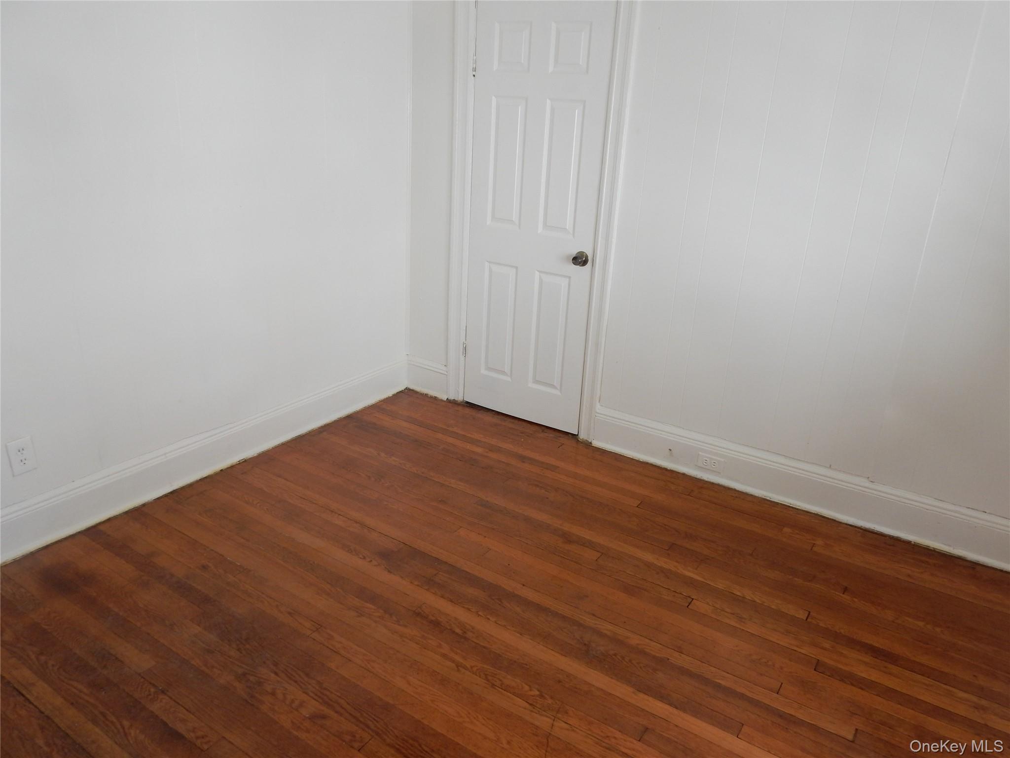 Empty room with reddish-brown hardwood floors, white paneled walls, and a six-panel door.