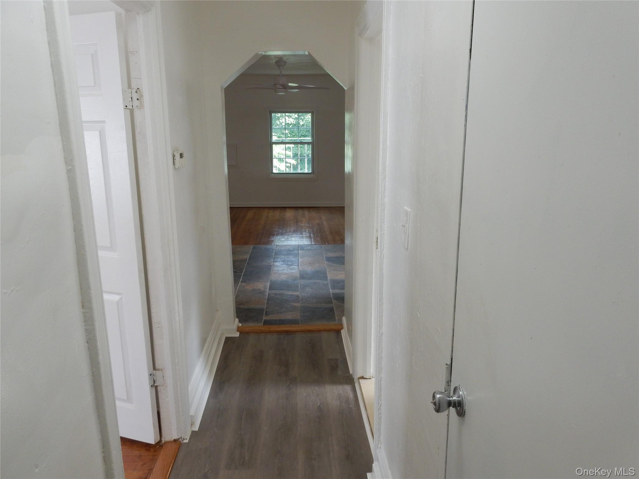 Hallway with gray flooring leads to an empty hardwood-floored room with a window and ceiling fan.