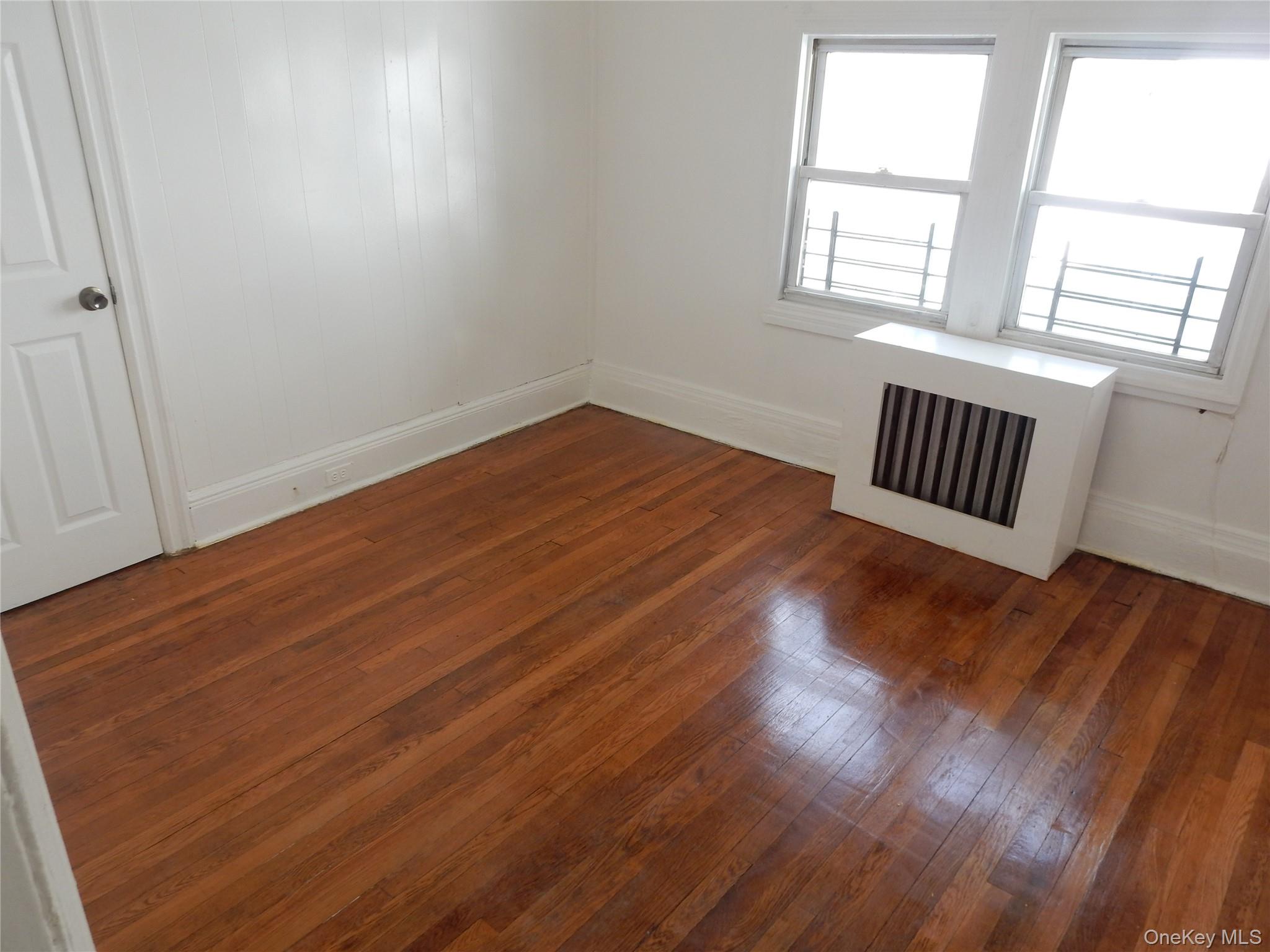 Empty room with polished hardwood floors, two windows, and a white radiator cover.