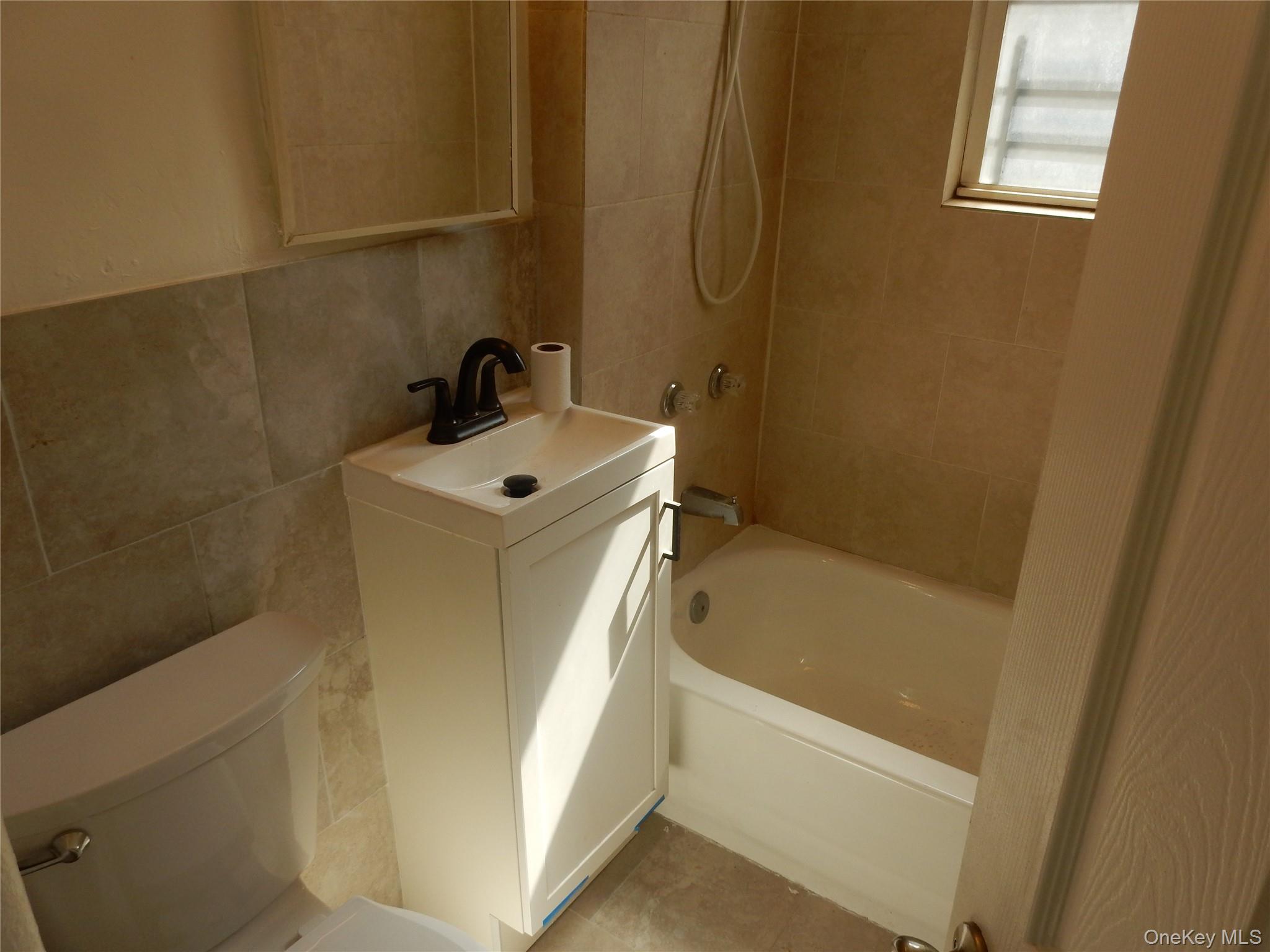 Bathroom with beige tile, a white vanity and black faucet, bathtub, shower, and partial view of a toilet.