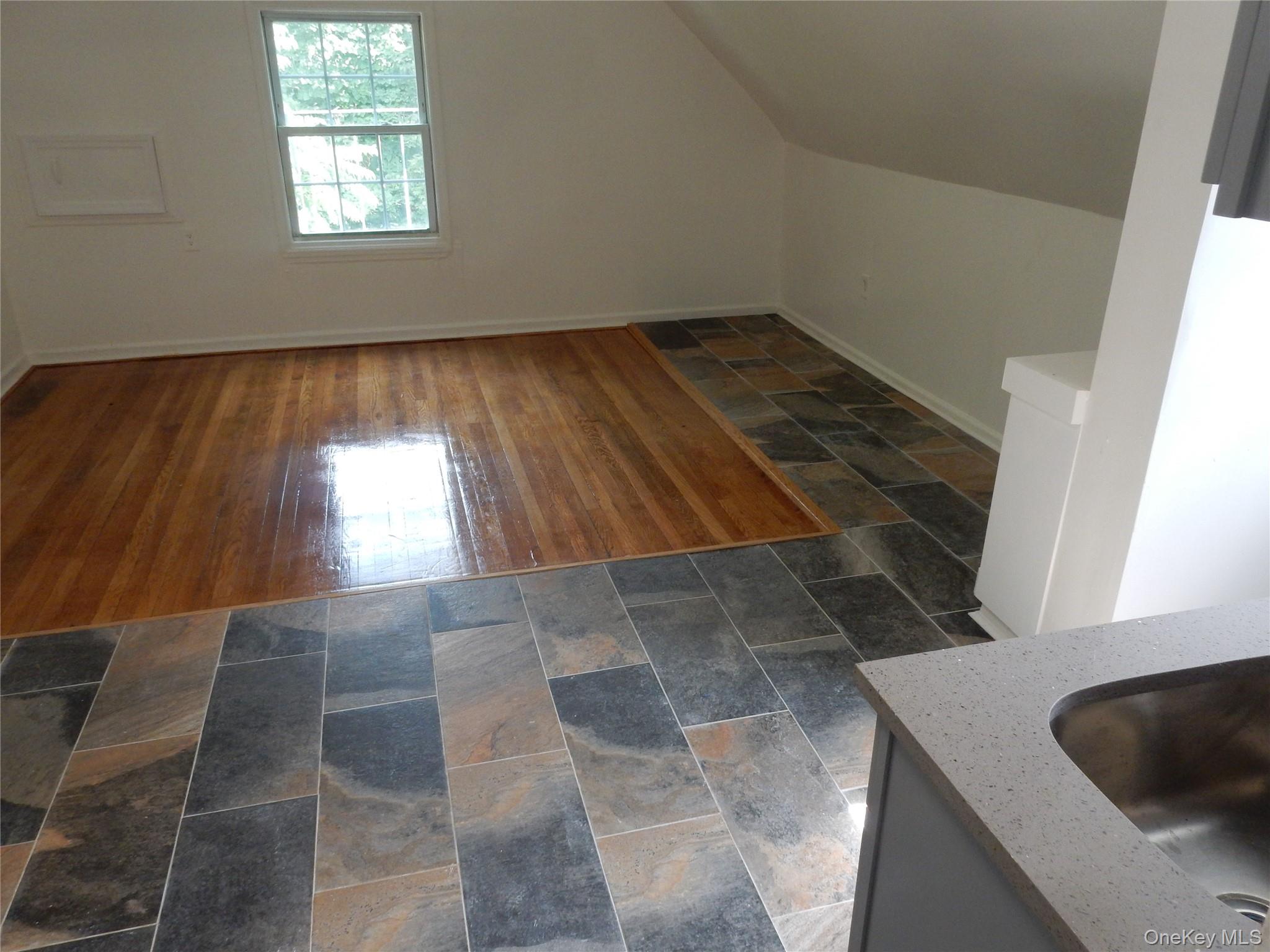 Empty kitchen with slate-look tile surrounding a polished hardwood floor inset and a window under a sloped ceiling.