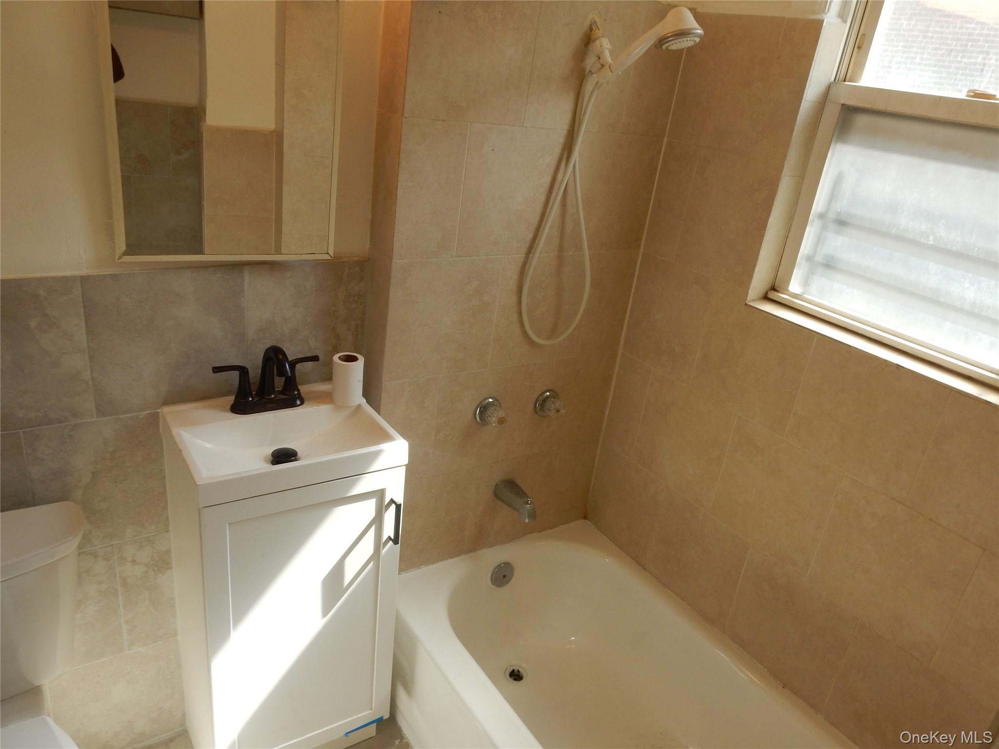 Bathroom with tiled walls, a bathtub and handheld shower, and a small vanity beside a window.