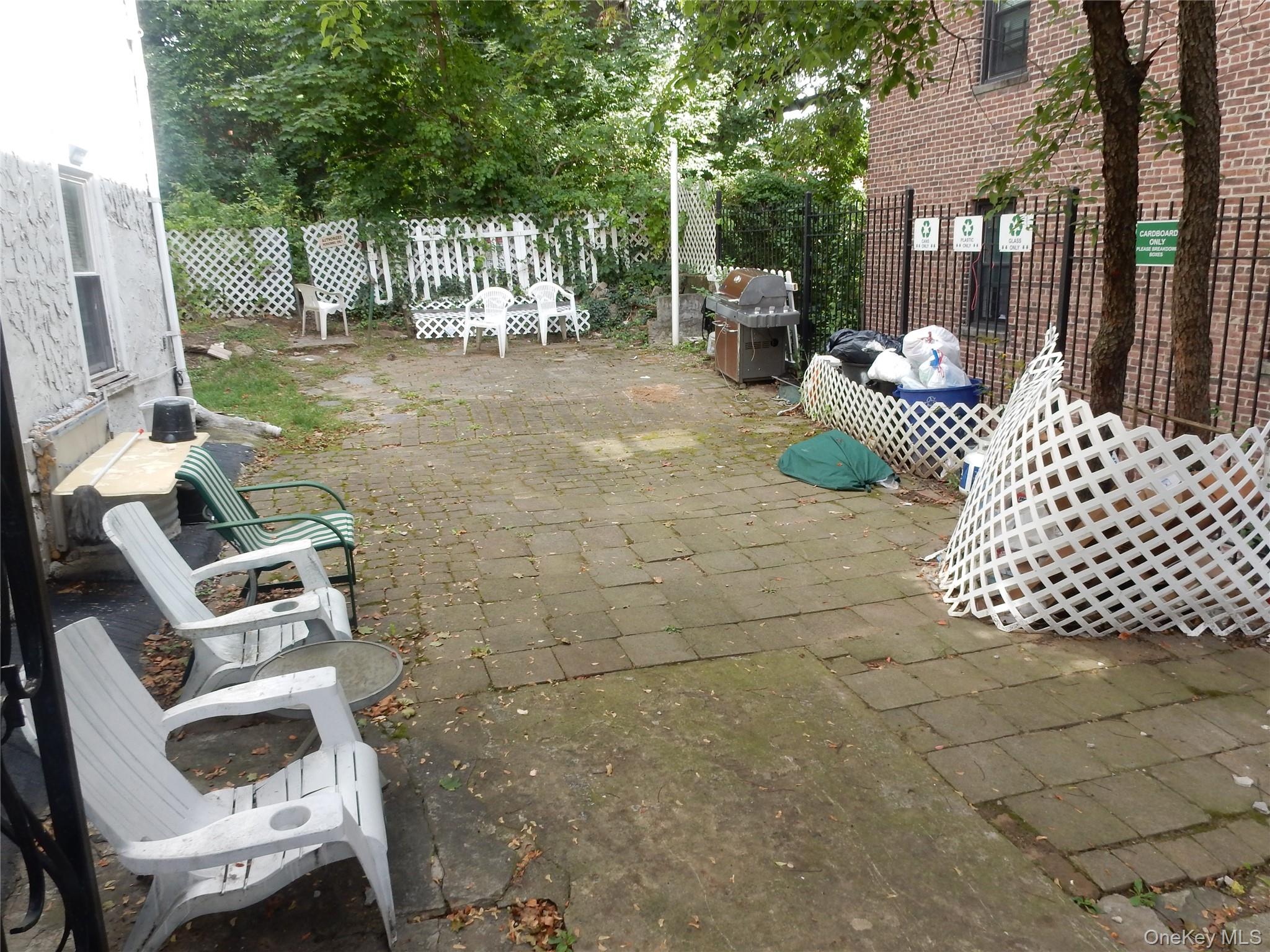 Paved patio with outdoor chairs, a barbecue grill, white lattice fencing, and leafy trees.