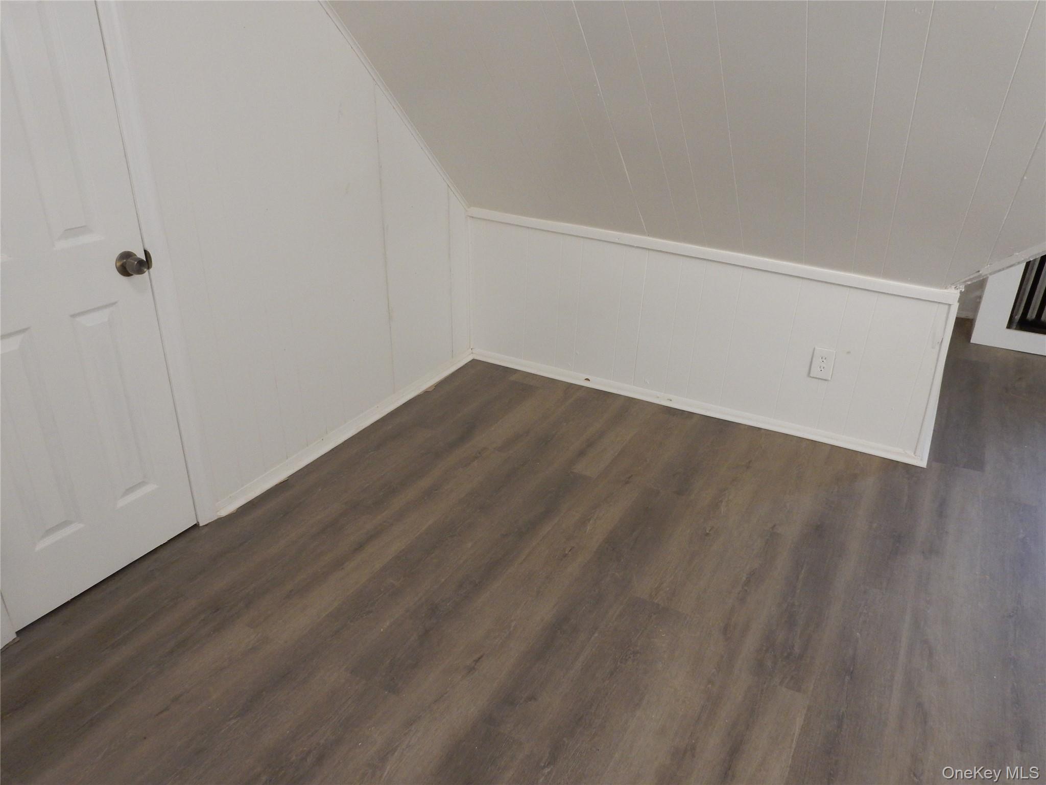 Empty room with a sloped ceiling, white paneled walls, and dark wood-look flooring.