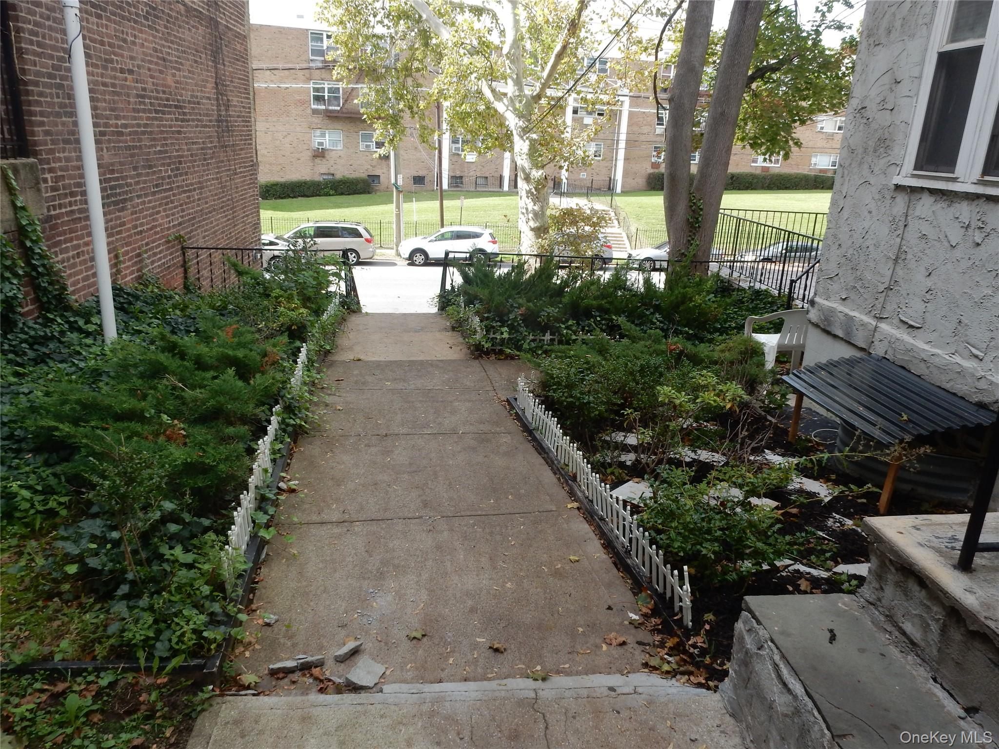 Concrete walkway through landscaped garden beds toward the street between buildings.