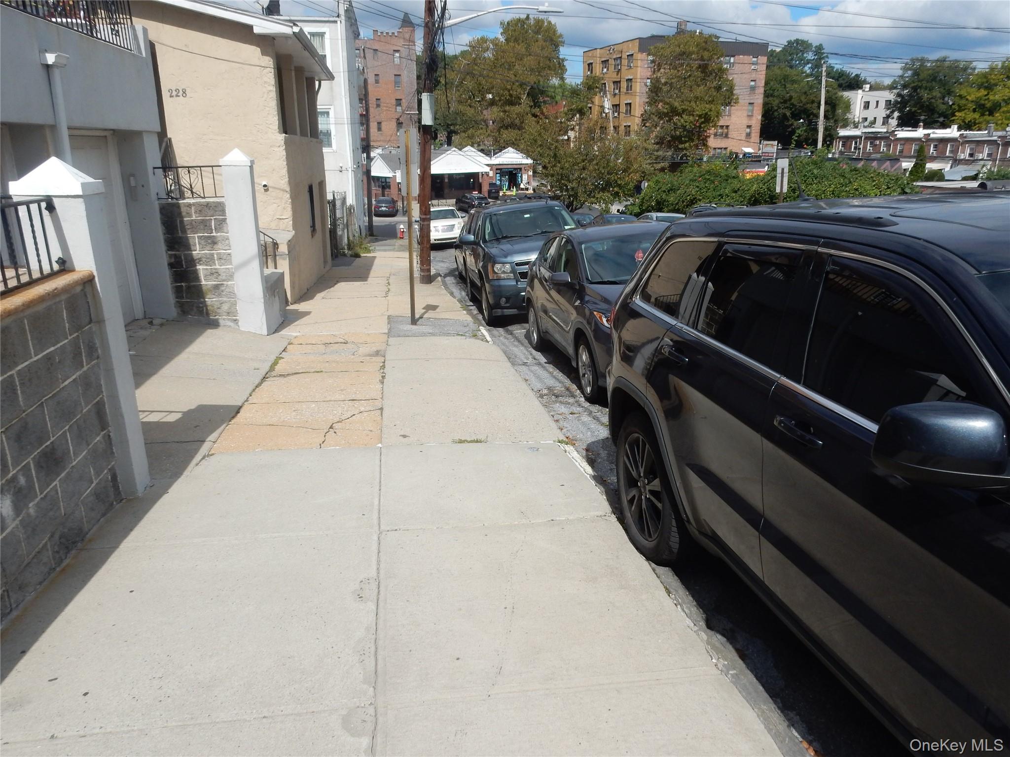 Sidewalk beside a light-colored building, with parked cars and apartment buildings along the street.