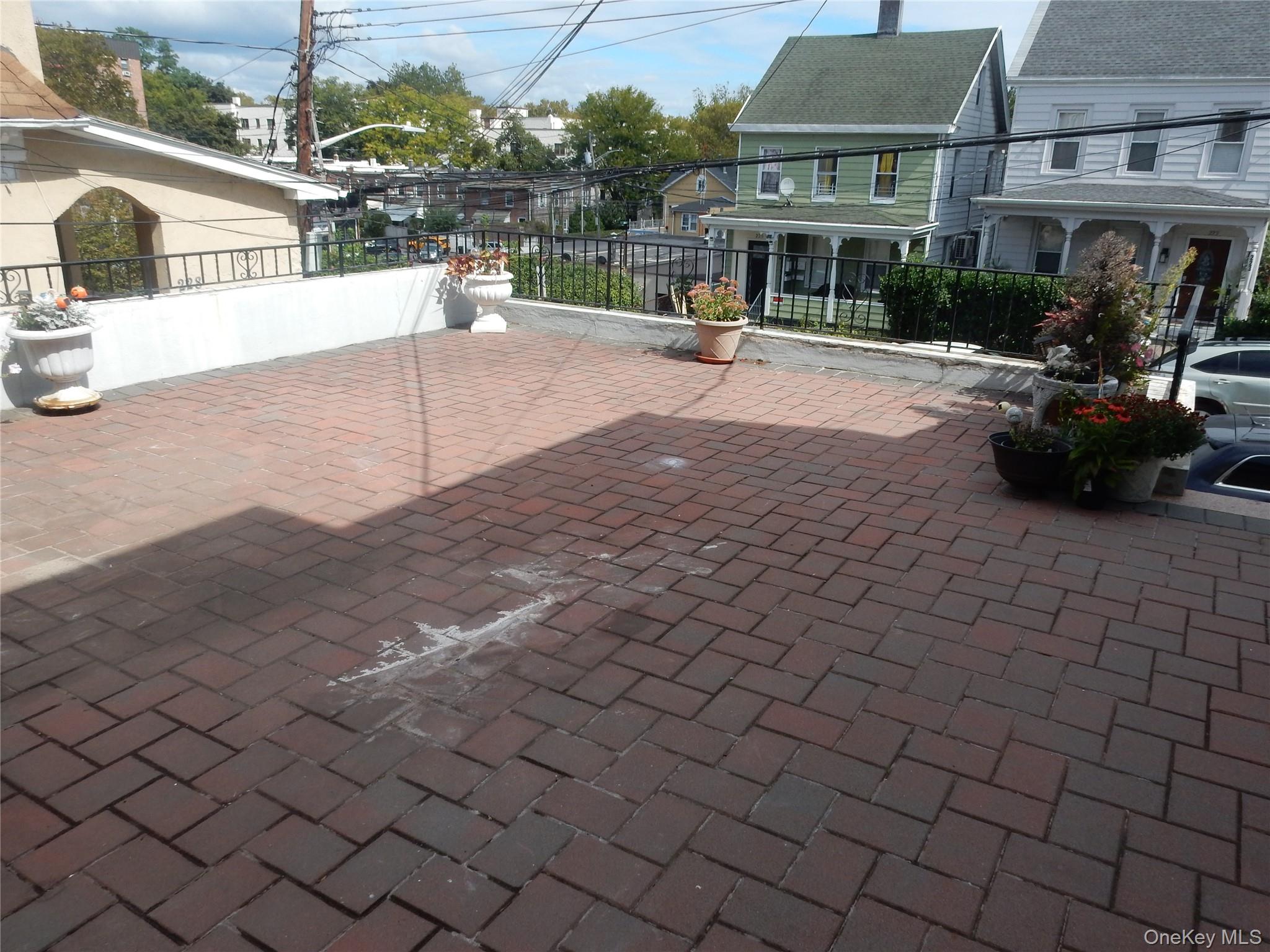 Rooftop terrace with reddish-brown pavers, black railing, and potted flowers overlooking neighboring houses.
