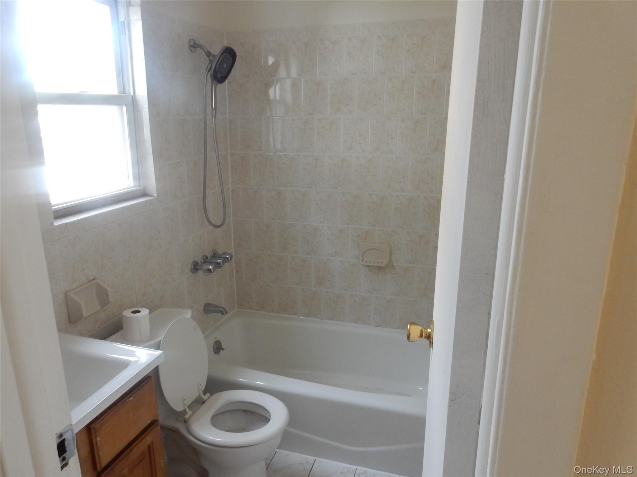 Bathroom with a tiled tub and shower, handheld showerhead, and window beside the tub.