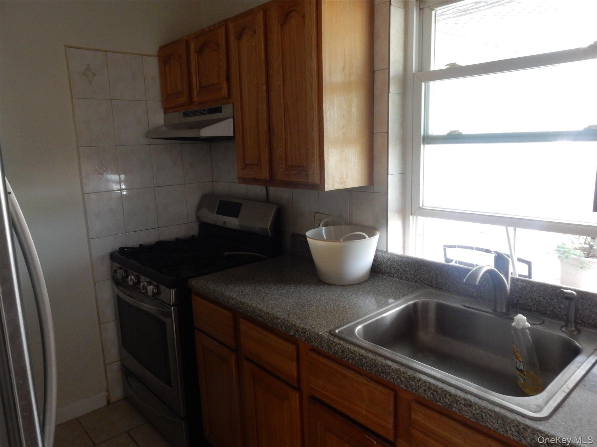 Kitchen with wood cabinets, granite counters, a stainless-steel sink, and gas range beside a bright window.