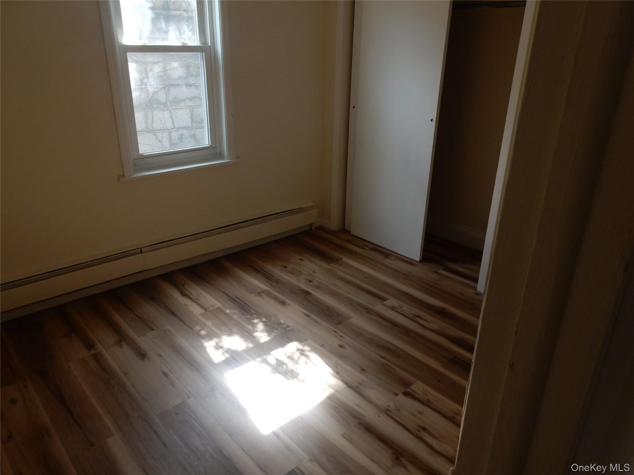 Empty room with wood plank floors, a window, baseboard heater, and sliding closet door.