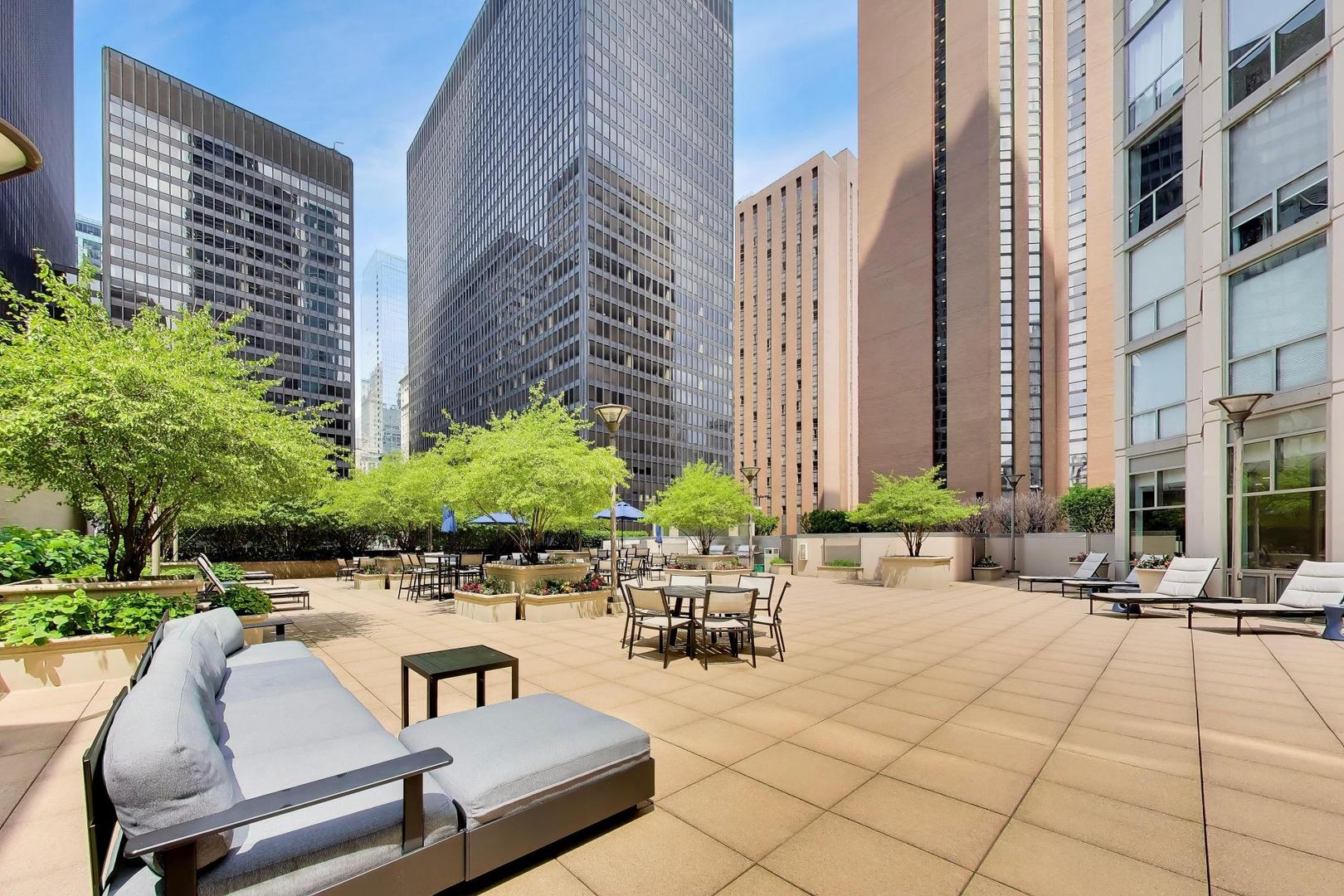 Rooftop terrace with lounge seating, dining tables, planters, and trees among downtown high-rises.