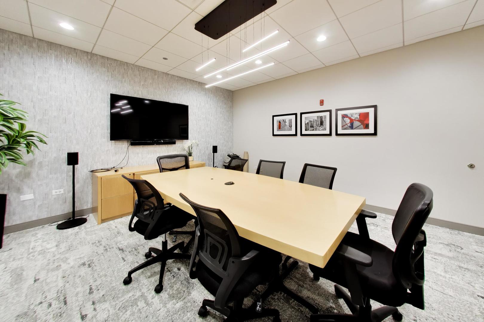 Furnished conference room with a long meeting table, black chairs, and wall-mounted television.