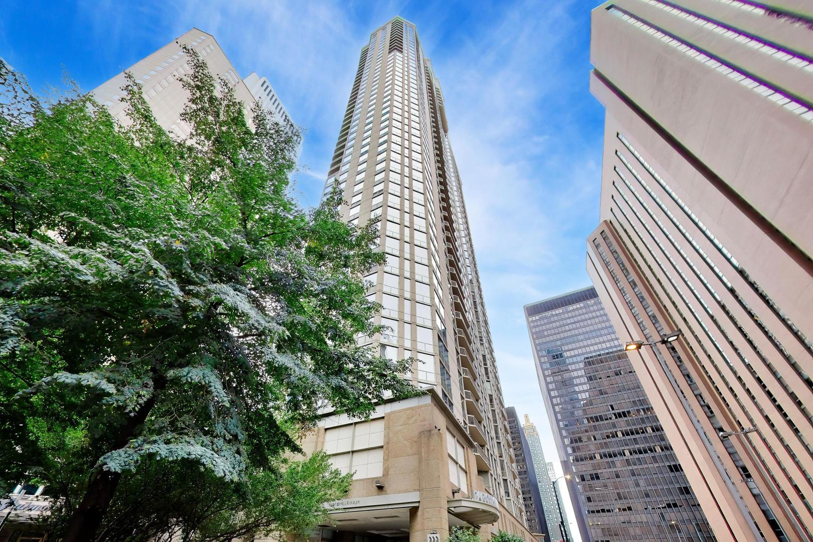 High-rise condominium tower seen from below among leafy trees and neighboring downtown buildings.